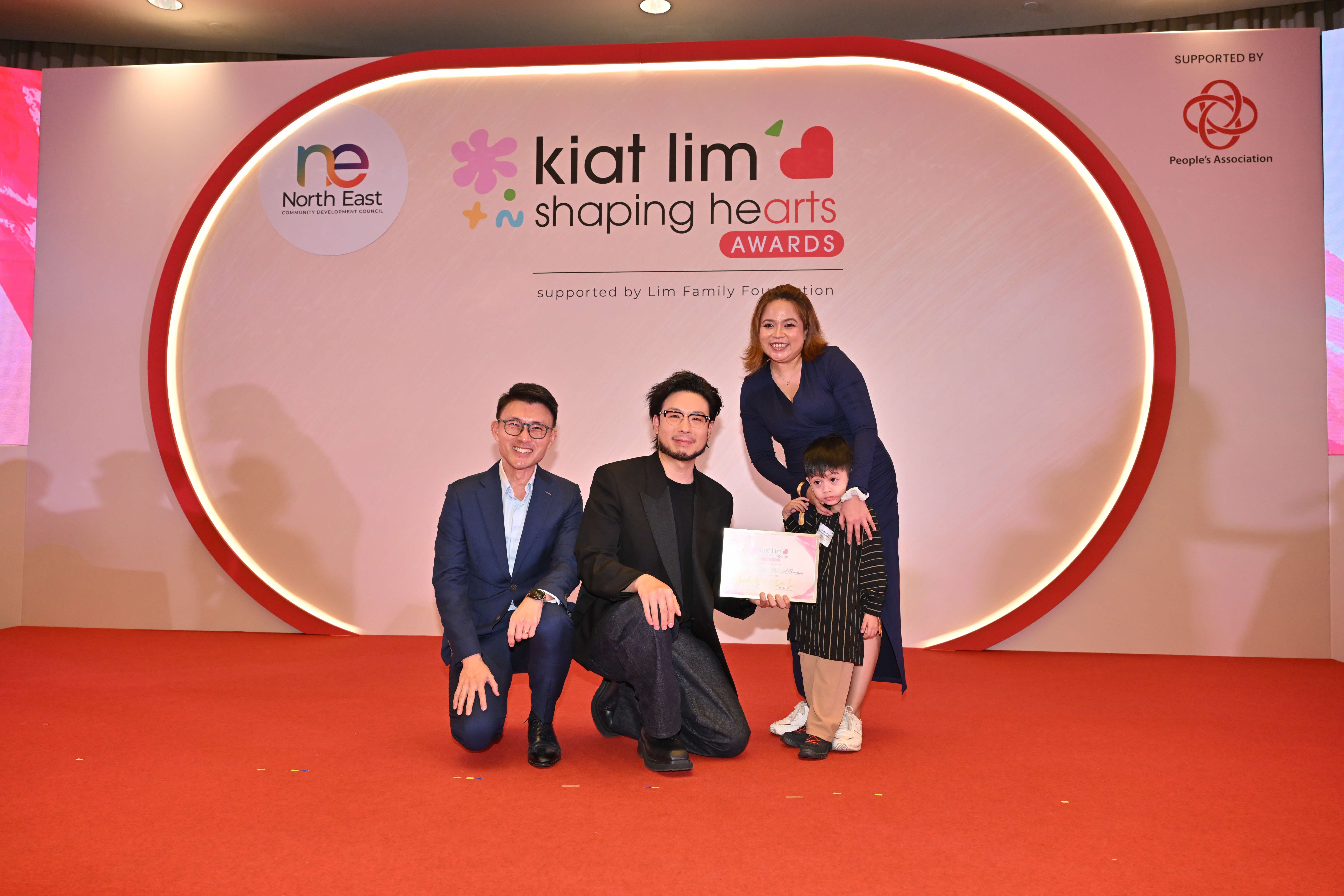 Award recipient standing on stage with the presenter, holding their certificate and posing for a group photo during the award ceremony, with a backdrop reading ‘Kiat Lim-Shaping Hearts Awards'.